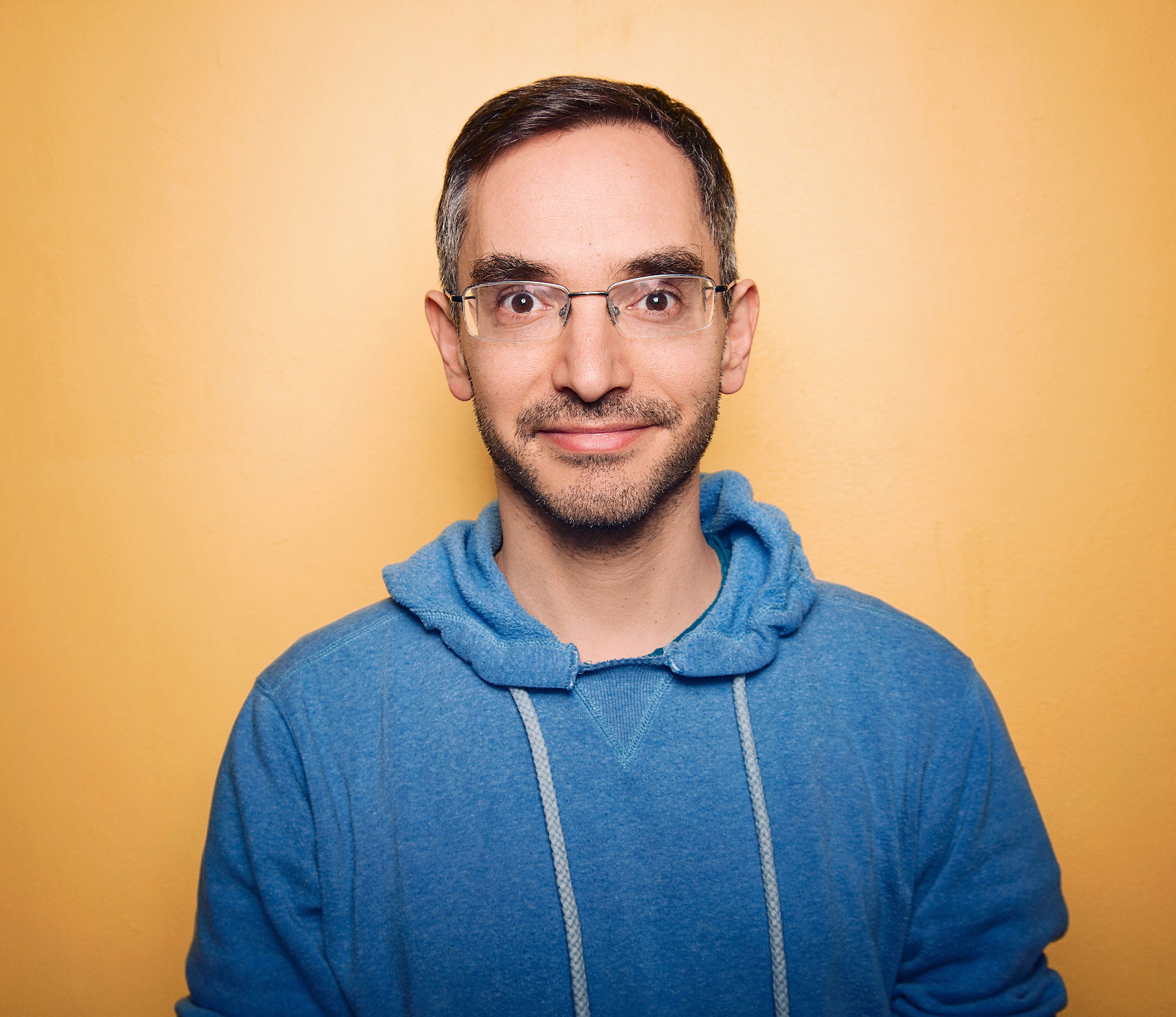 Myq Kaplan in a blue hoodie staring at the camera.