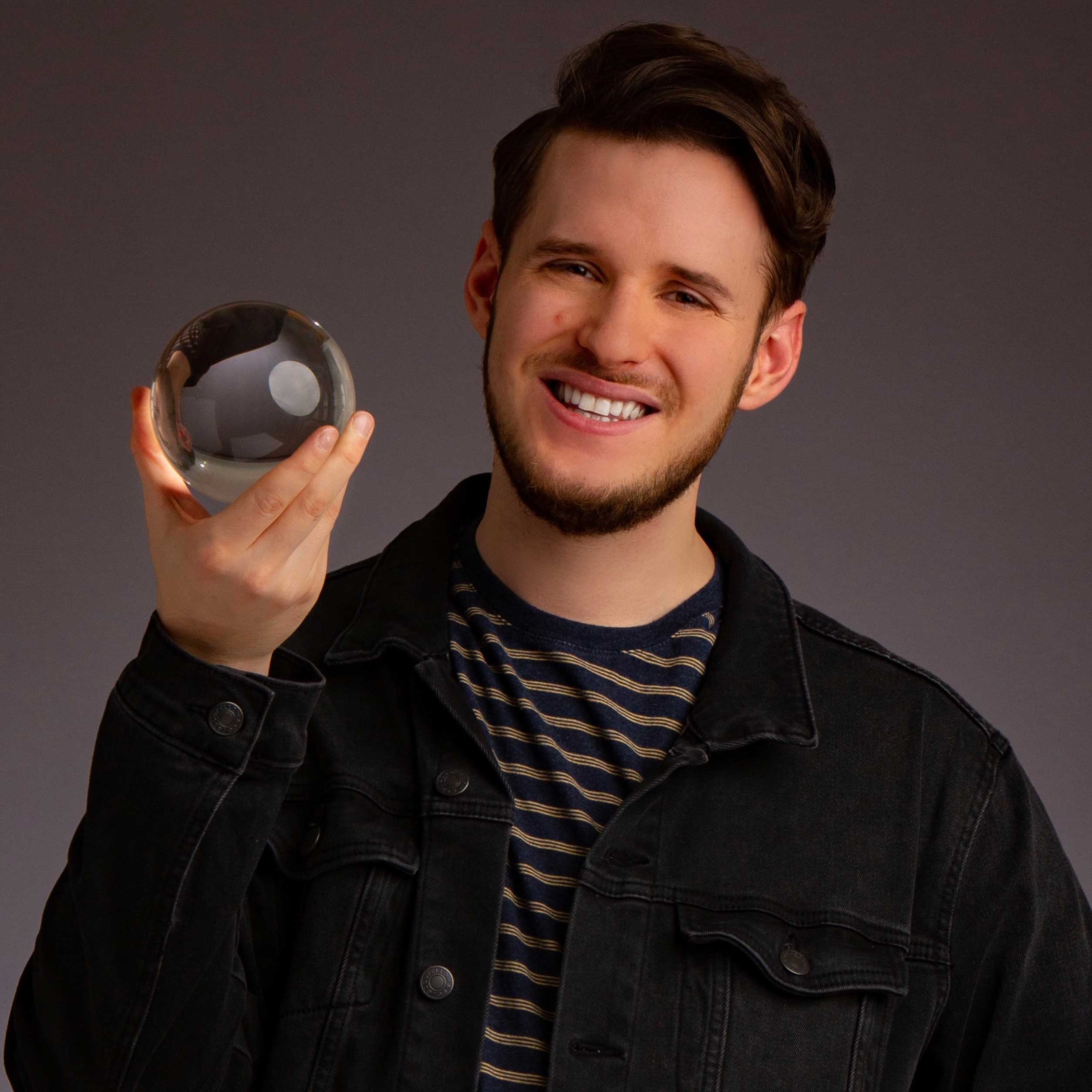 Jacob Mayfield smiling while holding crystal ball in hand wearing a black denim jacket over a striped shirt.