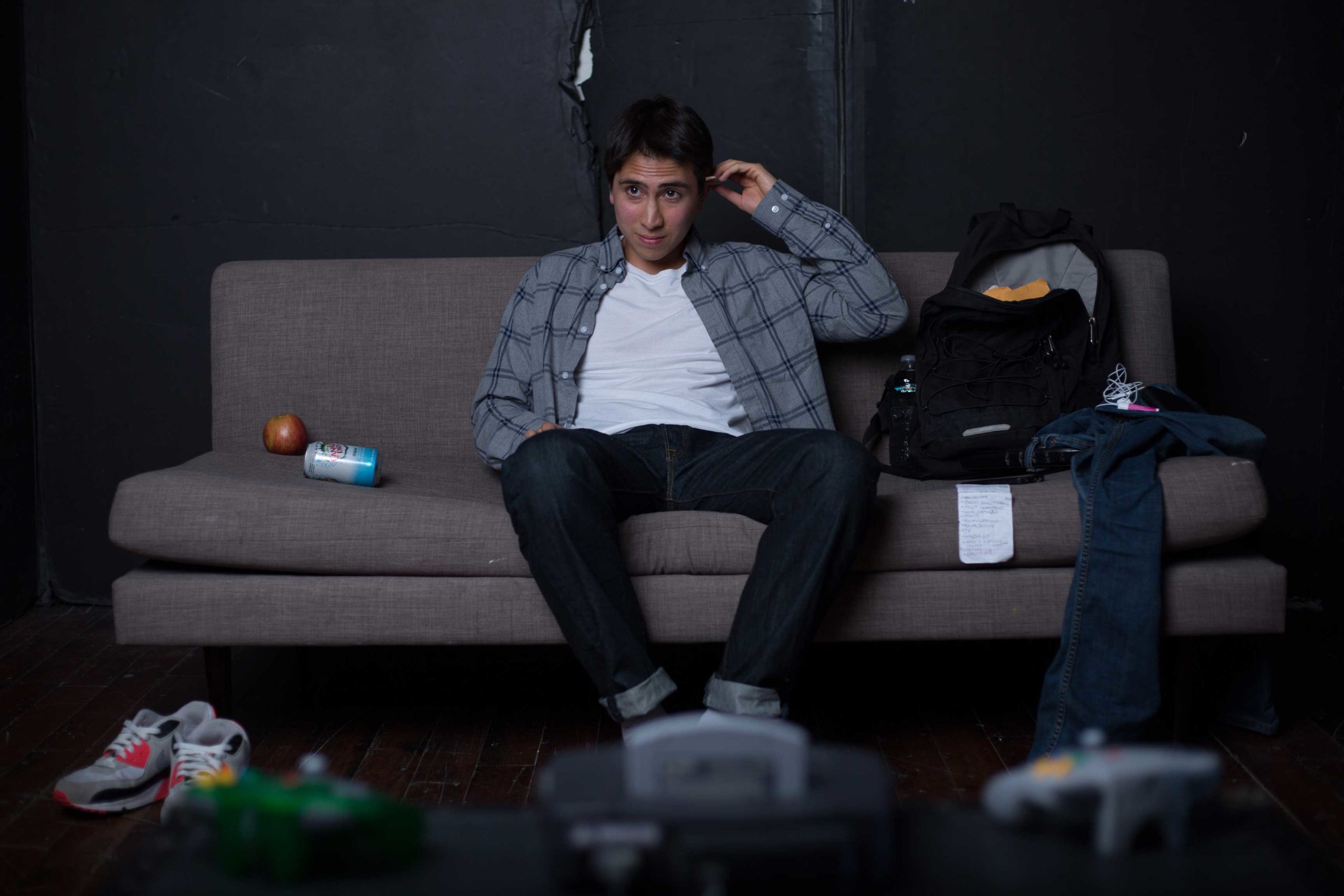 James Camacho sitting on a gray couch wearing a white t-shirt with a gray flannel and jeans.